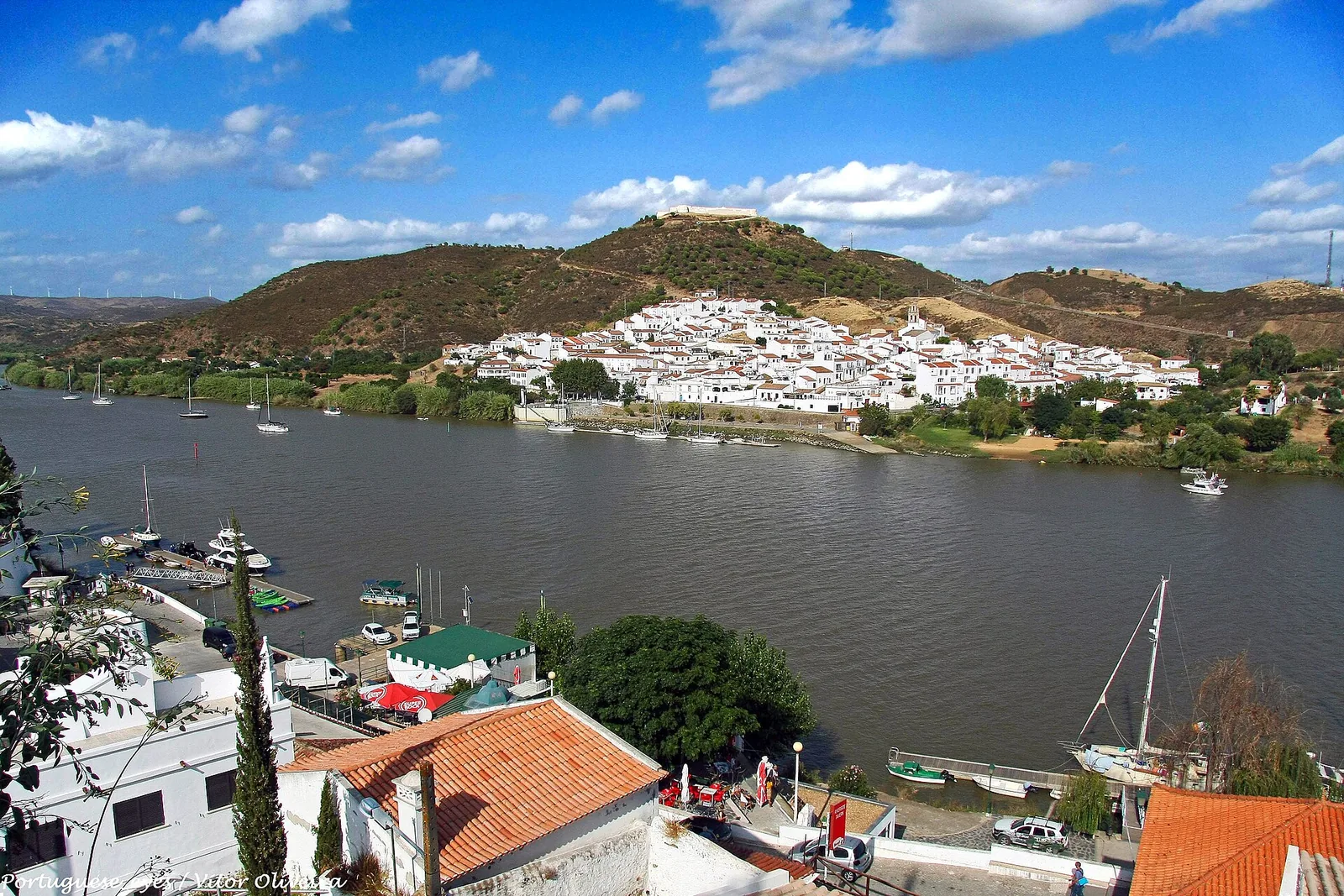 De kade van Alcoutim met aangemeerde zeilboten en terrasjes, en aan de overkant van de Guadiana het witte Sanlucar de Guadiana met zijn fort op de heuvel