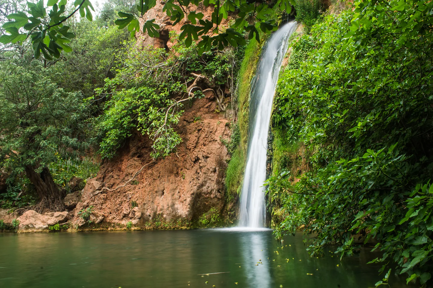 De waterval Queda do Vigário stort van een rode rotswand in een groene poel bij Alte