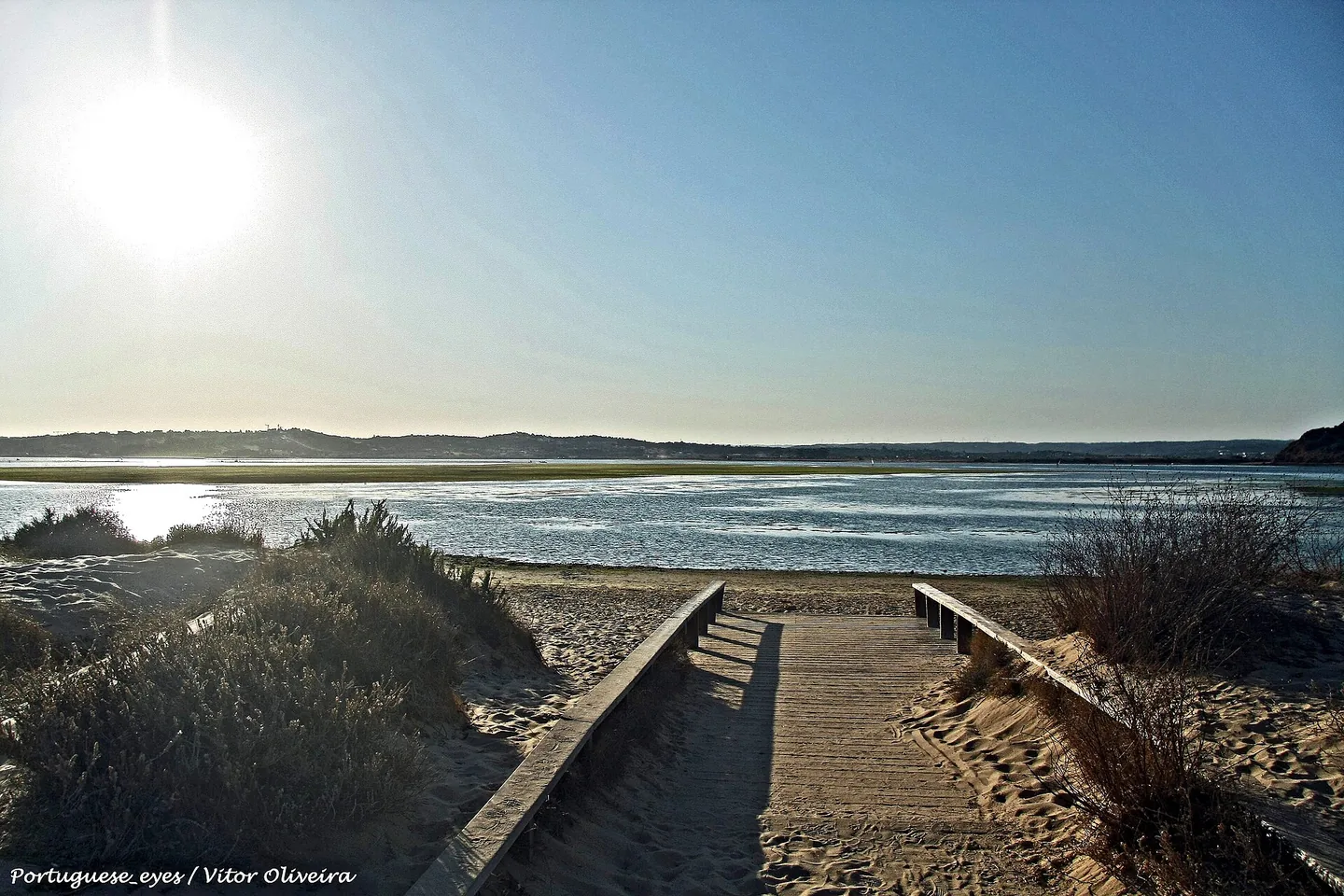 Een houten vlonderpad daalt door de duinen af naar de lagune van de Ria de Alvor, tegen de laagstaande zon