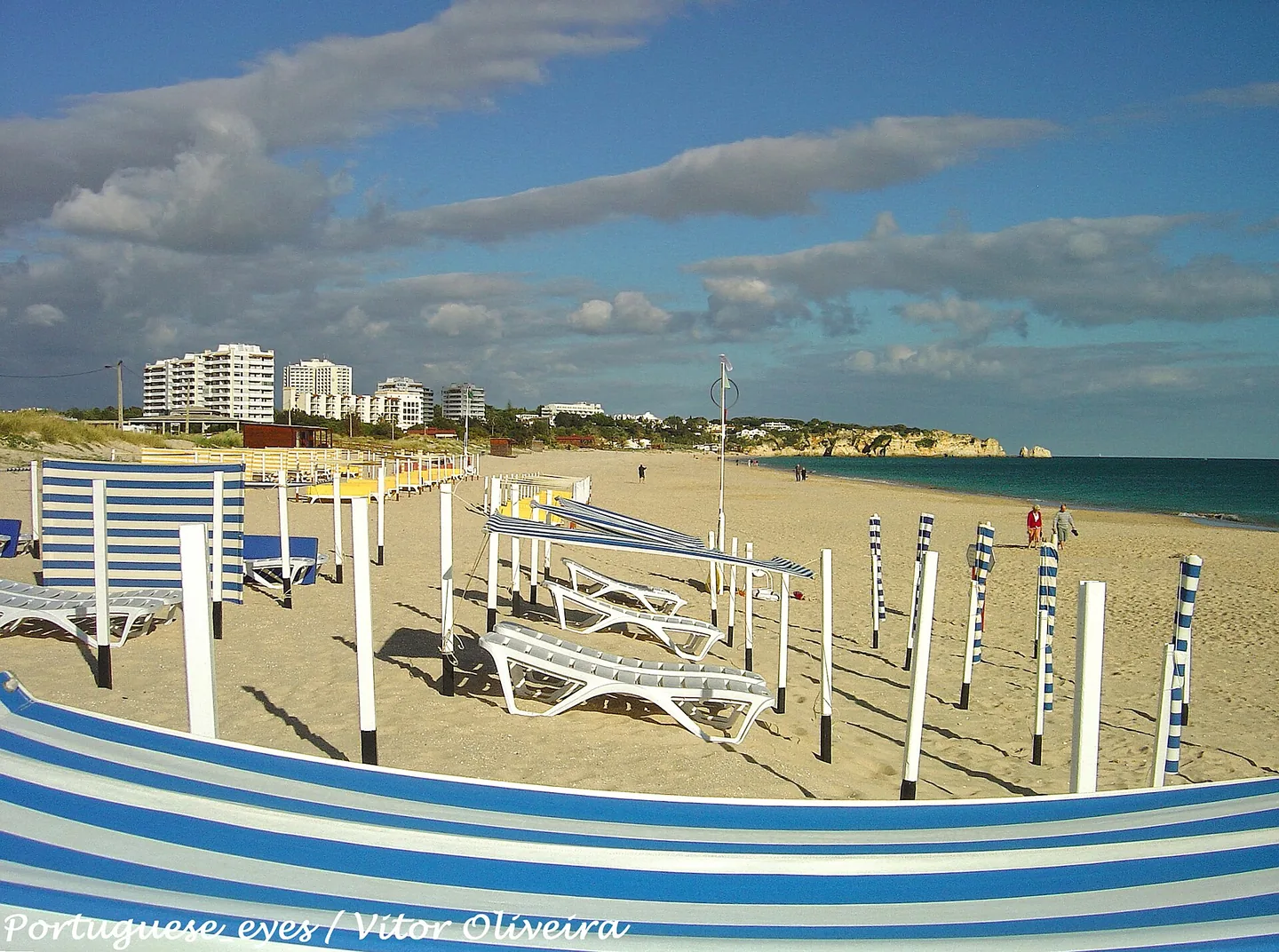 Ligbedden en gestreepte windschermen op het brede strand van Alvor, met appartementen op de achtergrond