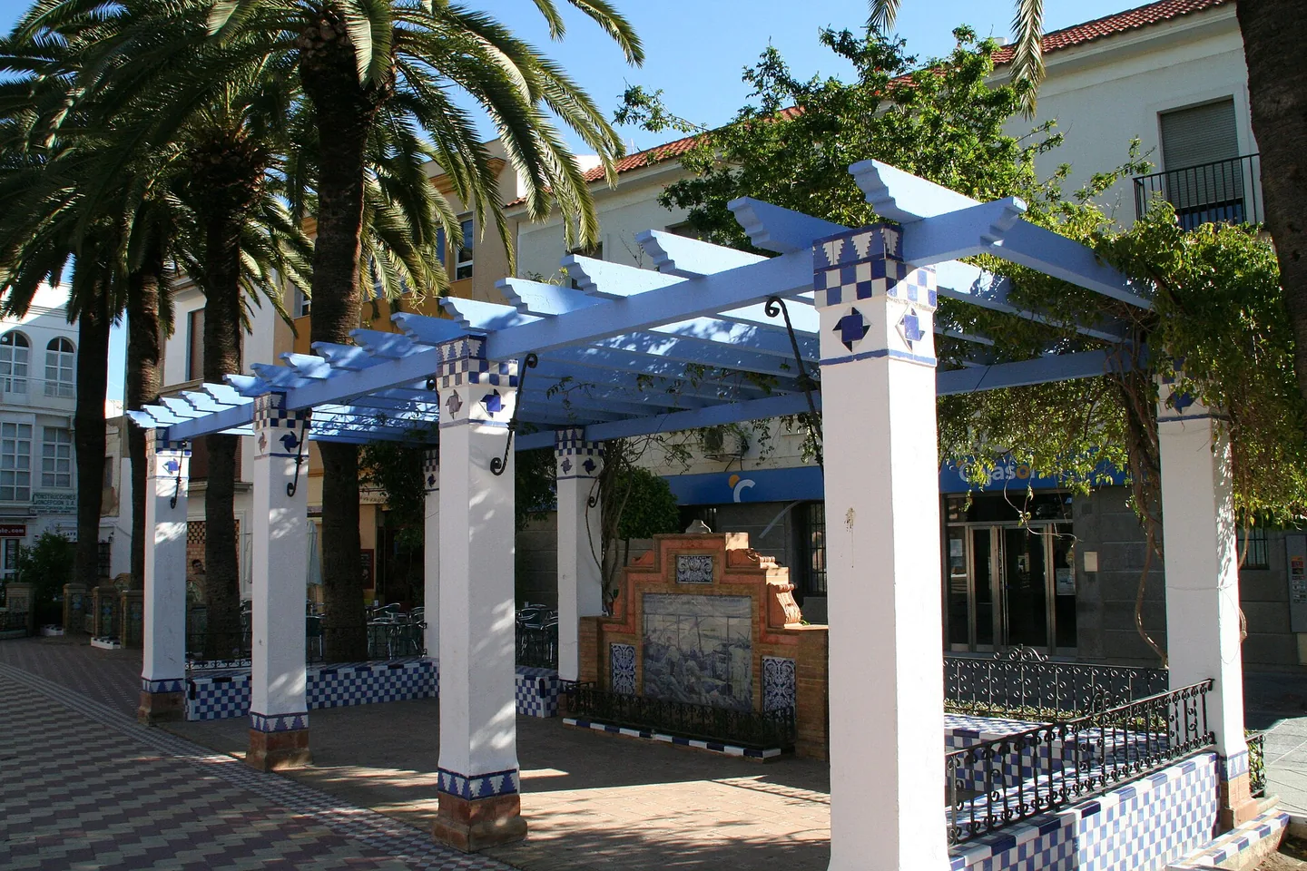 Witte pergola met lichtblauw geschilderde balken en blauw-witte azulejotegels op de Plaza de la Laguna in Ayamonte, met palmbomen en een terras erachter.