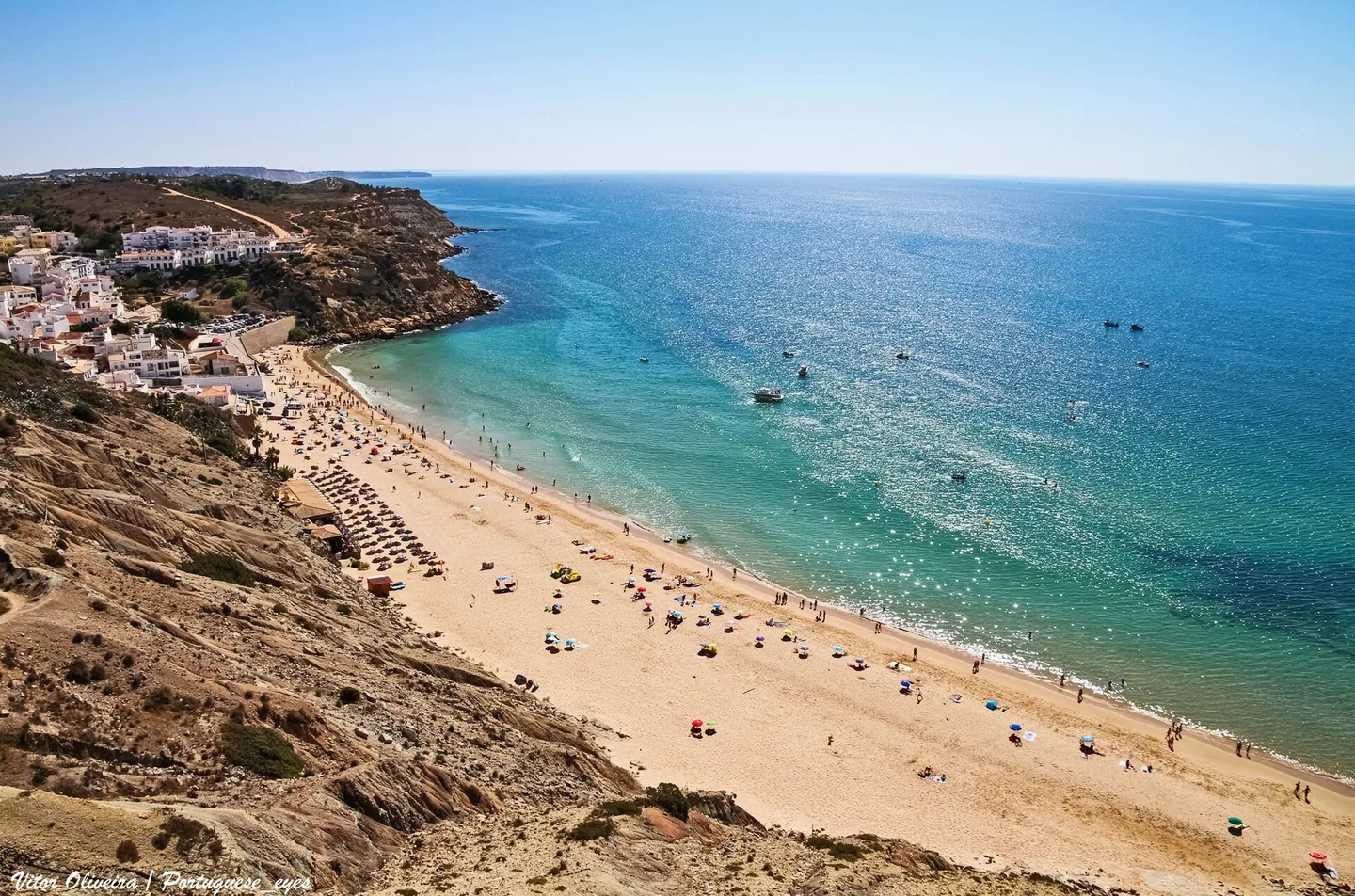 Het beschutte strand van Burgau met parasols en helderblauw water, tussen kliffen aan weerszijden