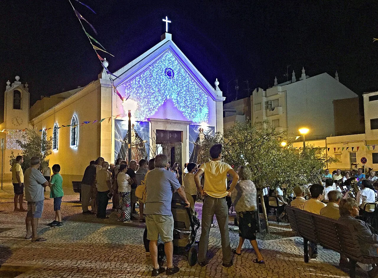 De verlichte parochiekerk van Monte Gordo tijdens een dorpsfeest, met bewoners op stoelen op het plein ervoor