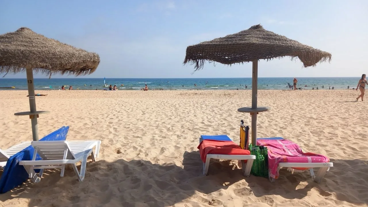 Twee rieten parasols met ligbedden op het brede zand van Praia de Monte Gordo, met de blauwe zee en badgasten op de achtergrond