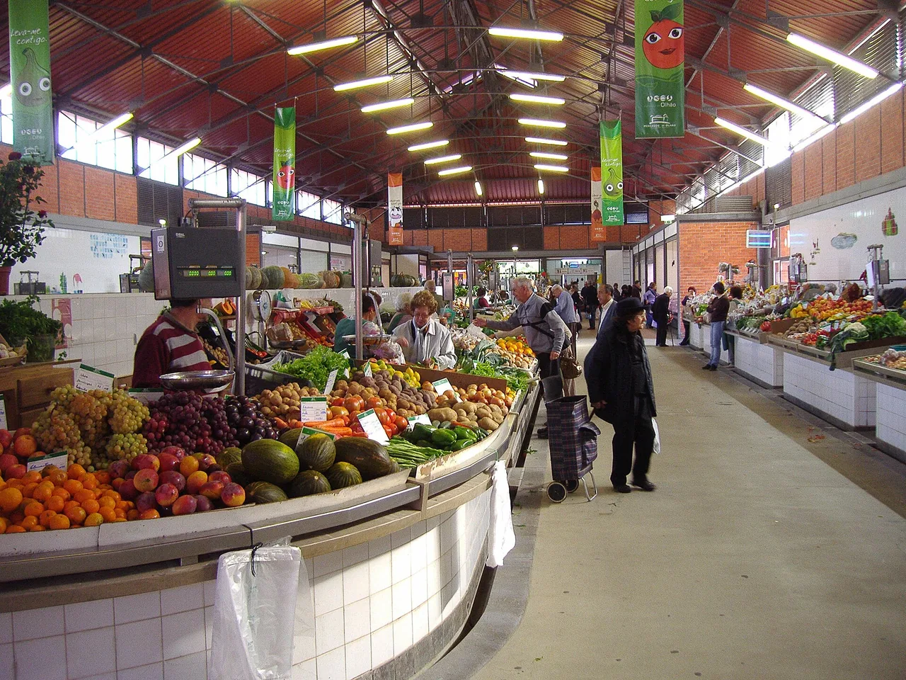 Kraam met vers fruit en groente in de overdekte markthal van Olhão: sinaasappels, druiven, meloenen en tomaten met prijskaartjes, en lokale klanten met boodschappentrolleys.