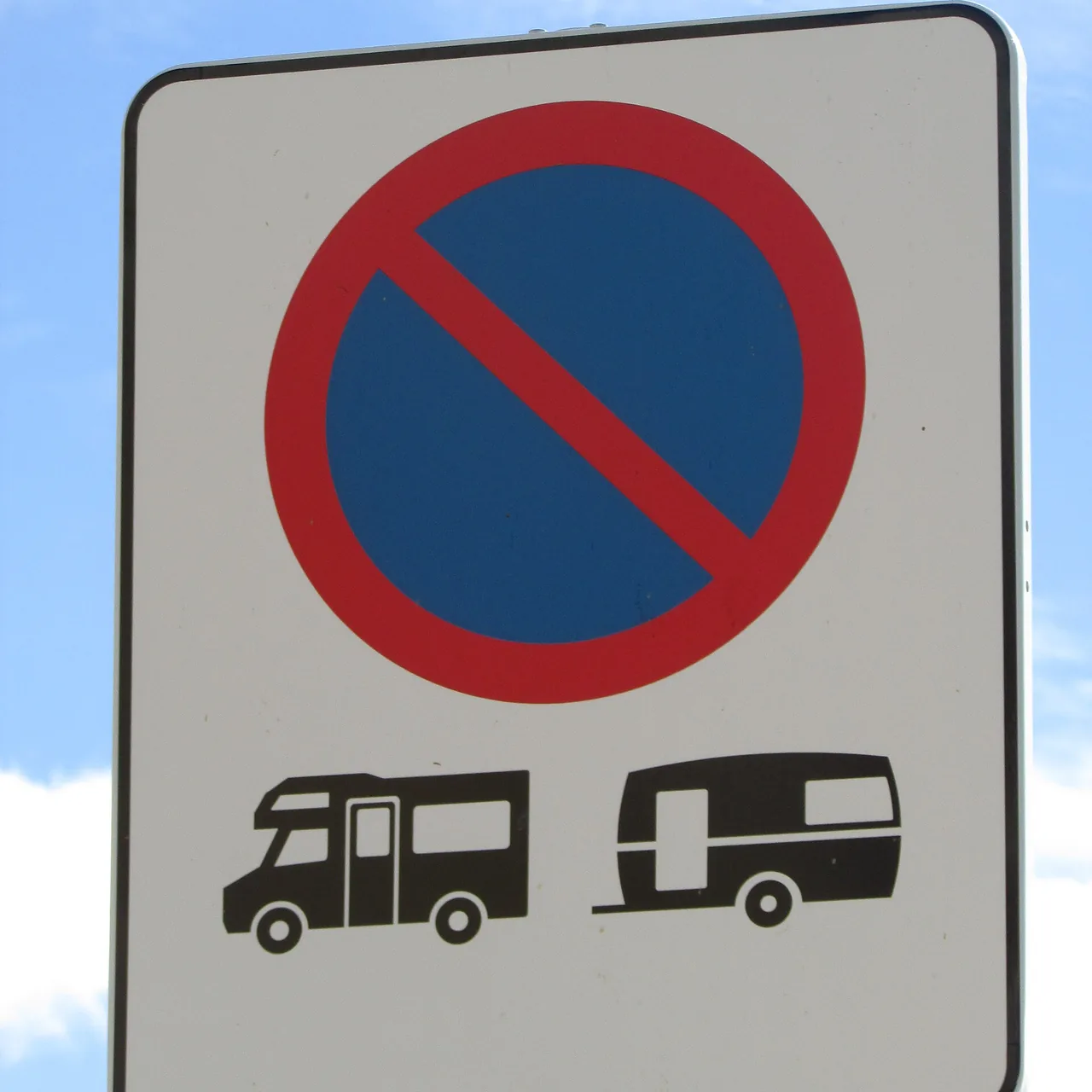 Verkeersbord met een rood-blauw parkeerverbodsteken en pictogrammen van een camper en een caravan, tegen een blauwe lucht met wolken