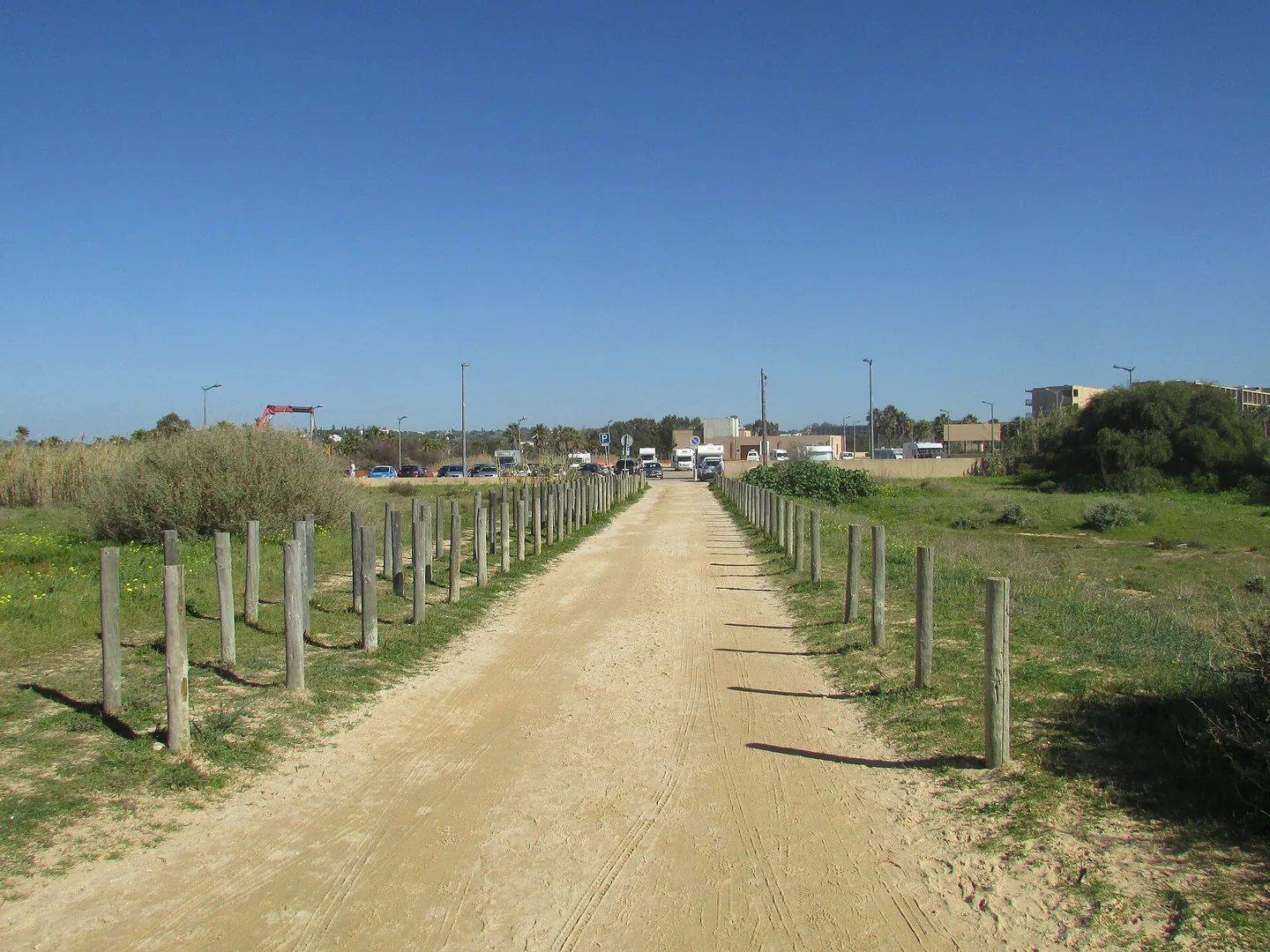 Zandpad met houten paaltjes naar de parkeerplaats achter Praia dos Salgados, met auto's en campers in de verte onder een strakblauwe lucht