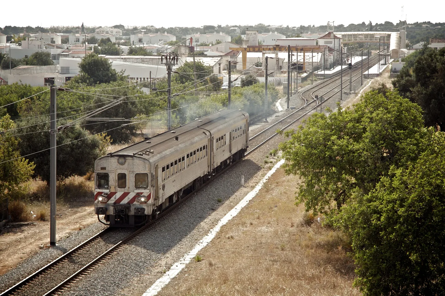 Een zilvergrijze regionale dieseltrein van CP met rood-witte strepen rijdt onder de bovenleiding naar station Albufeira-Ferreiras