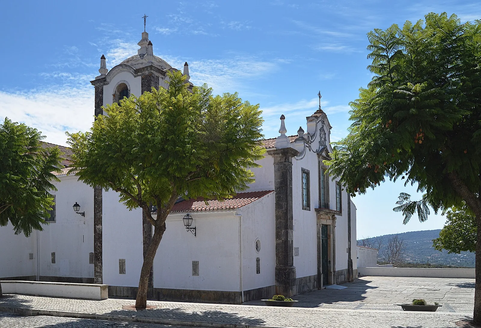 De witte Igreja Matriz van São Brás de Alportel met klokkentoren, geplaveid kerkplein en de heuvels van de barrocal erachter