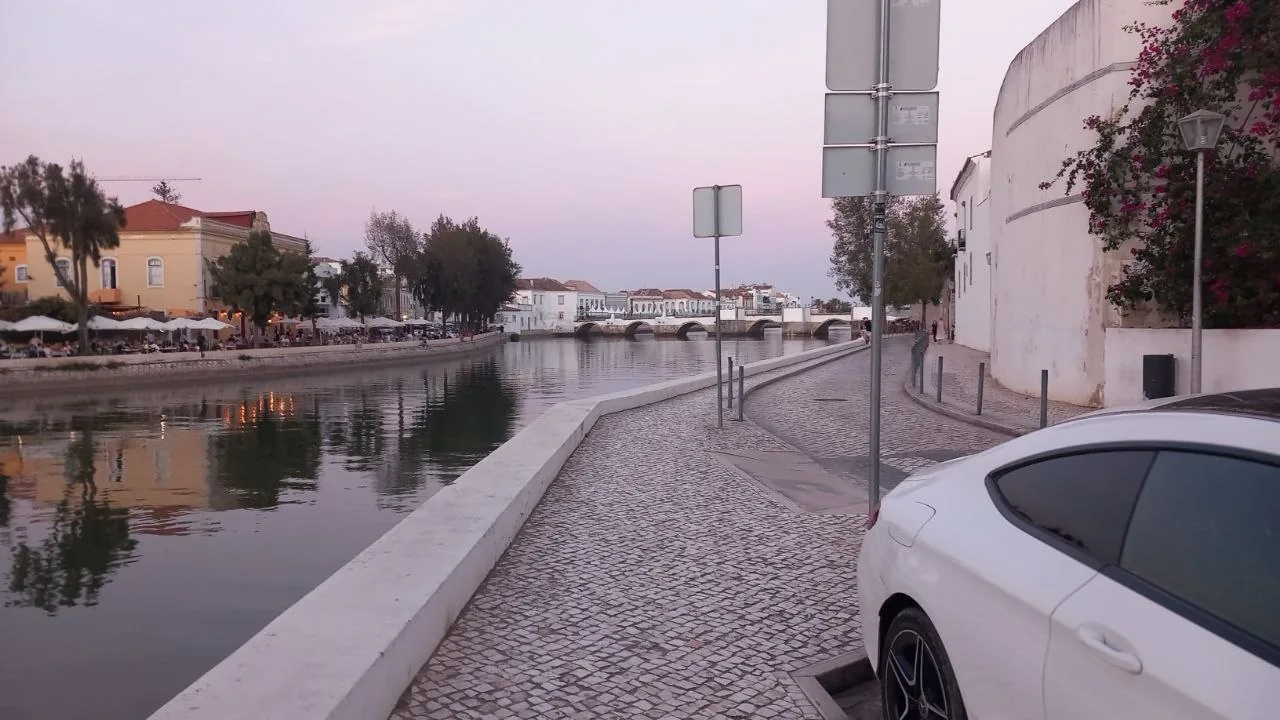 Dezelfde rivieroever van Tavira gezien vanaf de straat, met geparkeerde auto's langs de kade en terrassen aan de overkant