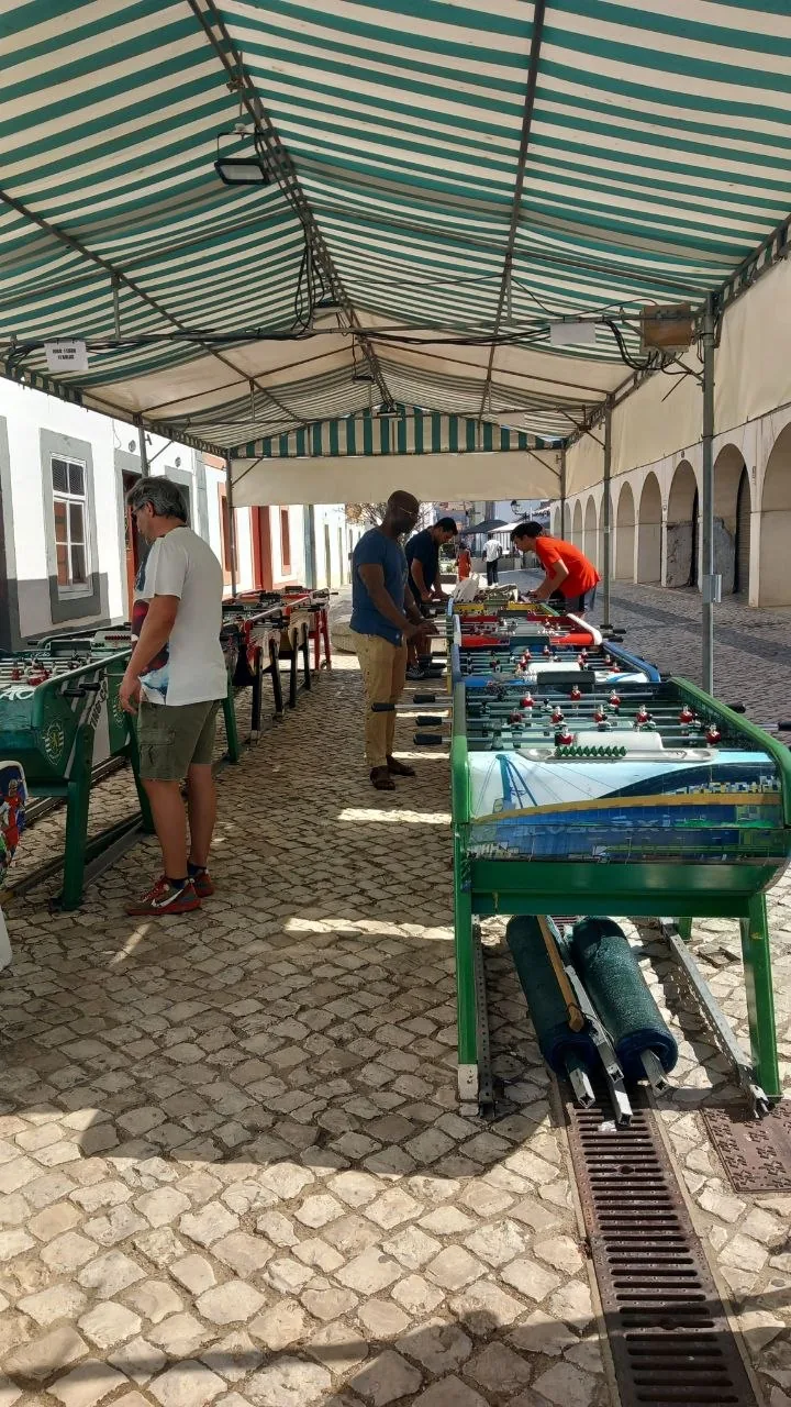 Een rij tafelvoetbaltafels onder een groen-wit gestreepte overkapping op een kasseienplein in Vila Real de Santo António